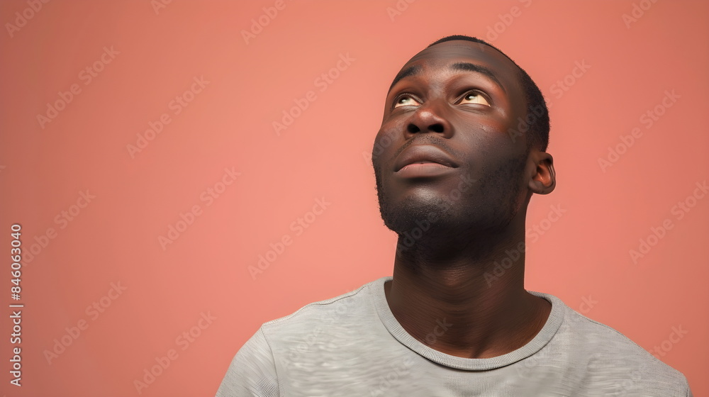 Fototapeta premium Studio shot of a black African American man, 20-27 years old, looking up. Thoughtful and interested, in a gray T-shirt, against a soft pink background.