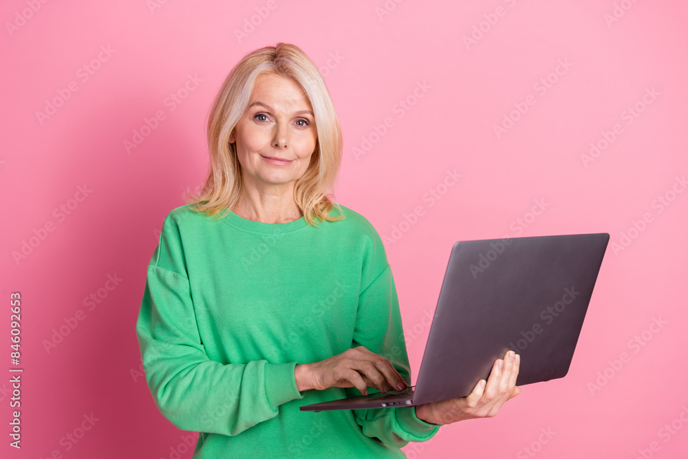 Photo portrait of lovely senior lady hold netbook dressed stylish green garment isolated on pink color background Photo portrait of lovely senior lady hold netbook dressed stylish green garment isolated on pink color background