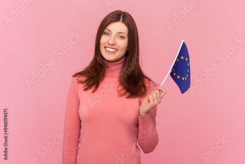 Woman with brown hair smiling broadly and showing flag of European Union, symbol of Europe, EU association and community, wearing rose turtleneck. Indoor studio shot isolated on pink background © khosrork