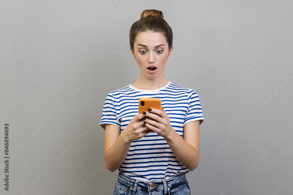 Φωτογραφία Portrait of beautiful astonished woman blogger wearing striped T-shirt using smart phone, looking at device screen, sees shocking content