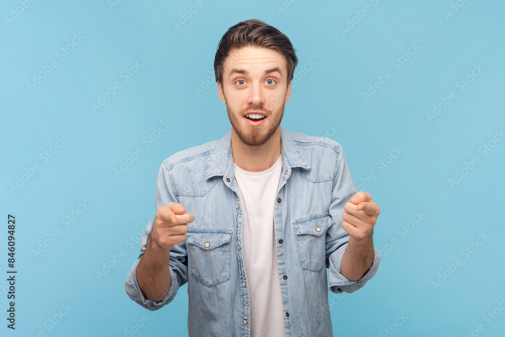 Portrait of astonished shocked surprised unshaven man points fore finger at camera, sees something unbelievable in front, wearing denim shirt. Indoor shot isolated on blue background. Portrait of astonished shocked surprised unshaven man points fore finger at camera, sees something unbelievable in front, wearing denim shirt. Indoor shot isolated on blue background.