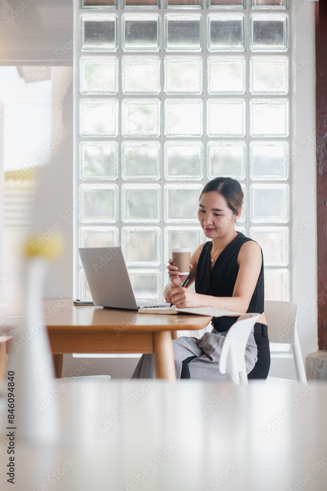 A young professional woman working on her laptop at a modern cafe, holding a coffee cup and smiling, with a glass block wall in the background. A young professional woman working on her laptop at a modern cafe, holding a coffee cup and smiling, with a glass block wall in the background.