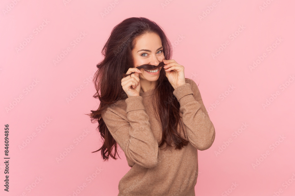 Portrait of joyful funny woman with wavy hair making mustache with her hair, looking at camera with positive emotions wearing wearing brown pullover. Indoor studio shot isolated on pink background Portrait of joyful funny woman with wavy hair making mustache with her hair, looking at camera with positive emotions wearing wearing brown pullover. Indoor studio shot isolated on pink background
