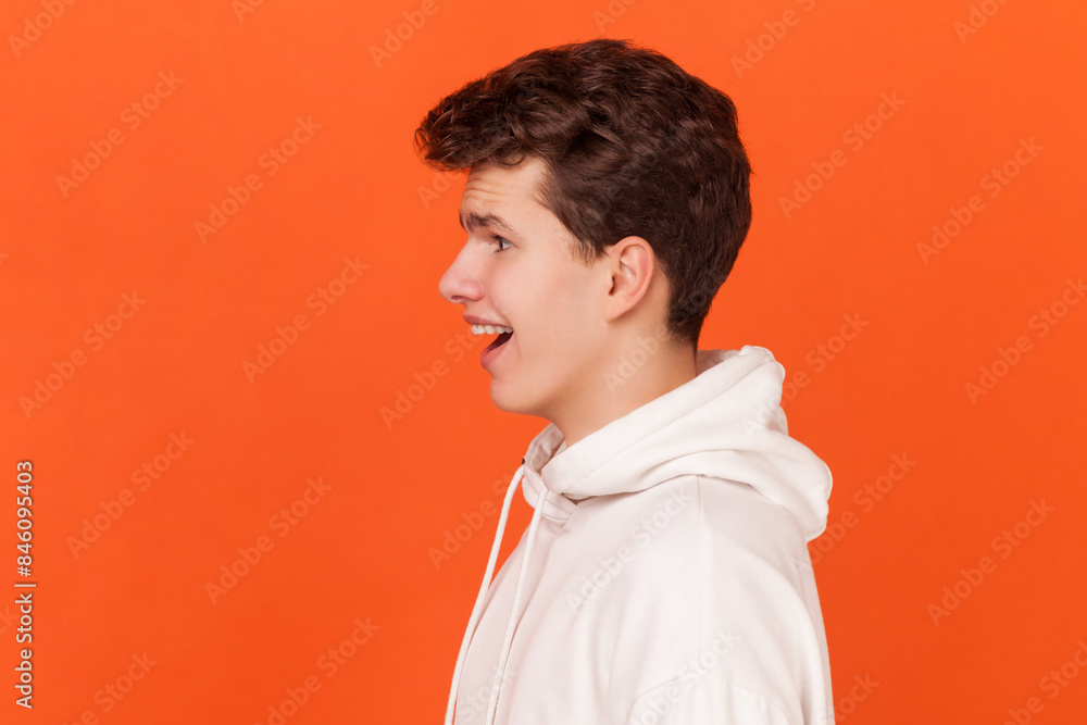 Side view portrait of astonished young handsome man wearing white hoodie looking at camera with open mouth, being pleasantly surprised by news. Indoor studio shot isolated on orange background. Side view portrait of astonished young handsome man wearing white hoodie looking at camera with open mouth, being pleasantly surprised by news. Indoor studio shot isolated on orange background.