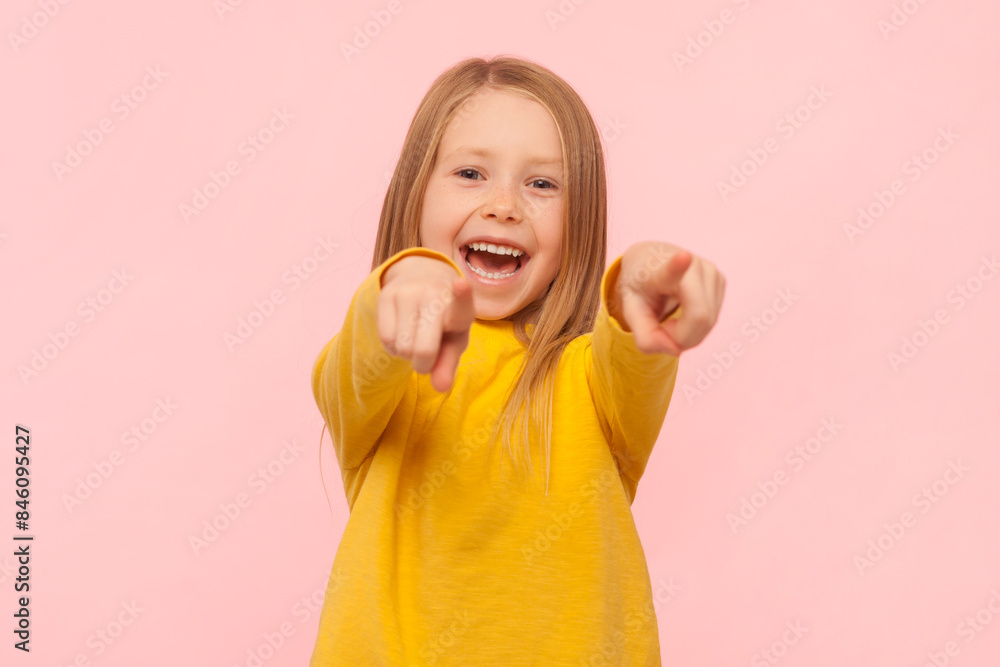 Portrait of satisfied excited smiling blonde little girl pointing at camera with both hands, choosing you to play together, wearing yellow jumper. Indoor studio shot isolated on pink background. Portrait of satisfied excited smiling blonde little girl pointing at camera with both hands, choosing you to play together, wearing yellow jumper. Indoor studio shot isolated on pink background.