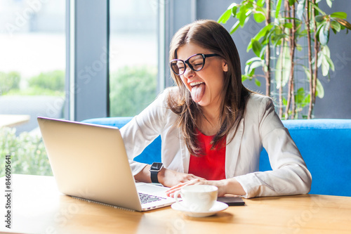Portrait of playful funny woman working on laptop showing tongue out having fun while having online meeting, wearing jacket and re shirt. Indoor shot, cafe or office background.