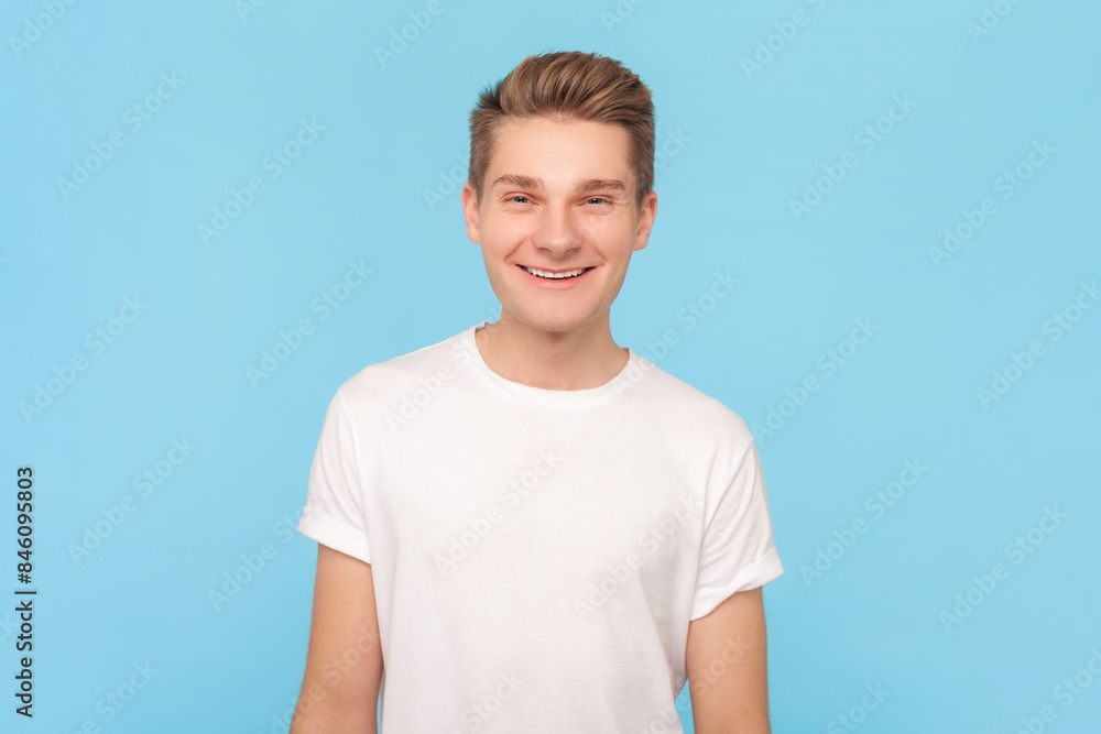 Portrait of smiling cheerful young man wearing white t-shirt expressing joy and happiness, smiling to camera, being in good mood. Indoor studio shot isolated on blue background Portrait of smiling cheerful young man wearing white t-shirt expressing joy and happiness, smiling to camera, being in good mood. Indoor studio shot isolated on blue background
