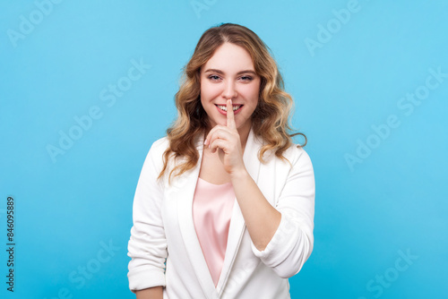 Portrait of smiling cheerful blond woman holding finger near mouth keeping secret, looking at camera with positive expression, wearing white shirt. Indoor studio shot isolated on blue background.
