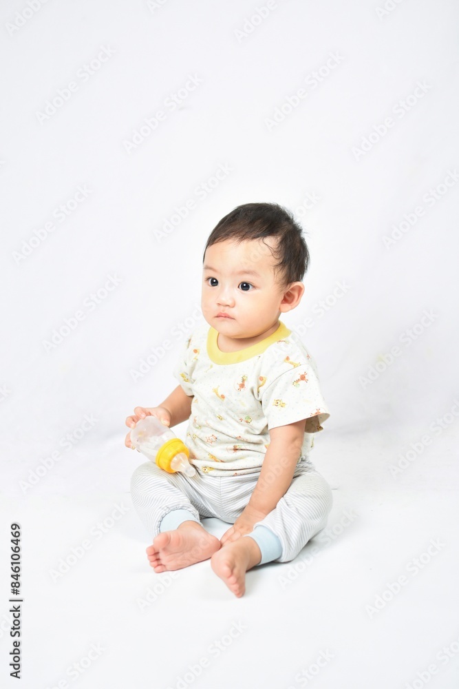 A cute baby with a sad expression, sitting and holding a milk bottle filled with water. The baby wears a light-colored outfit with animal prints, seated on a white background. A cute baby with a sad expression, sitting and holding a milk bottle filled with water. The baby wears a light-colored outfit with animal prints, seated on a white background.
