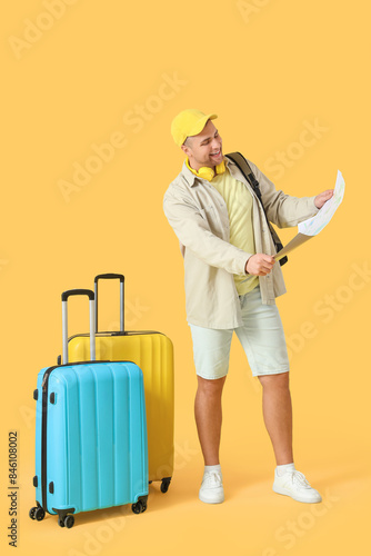 Male tourist with suitcases looking at map on yellow background