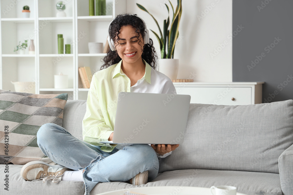 Young African-American woman in headphones using laptop on sofa at home Young African-American woman in headphones using laptop on sofa at home