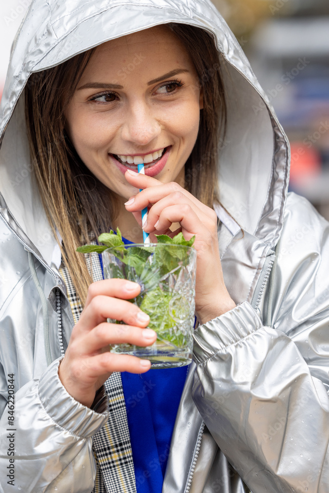 Φωτογραφία A young woman in a silver hooded jacket happily drinking a mint beverage outdoor