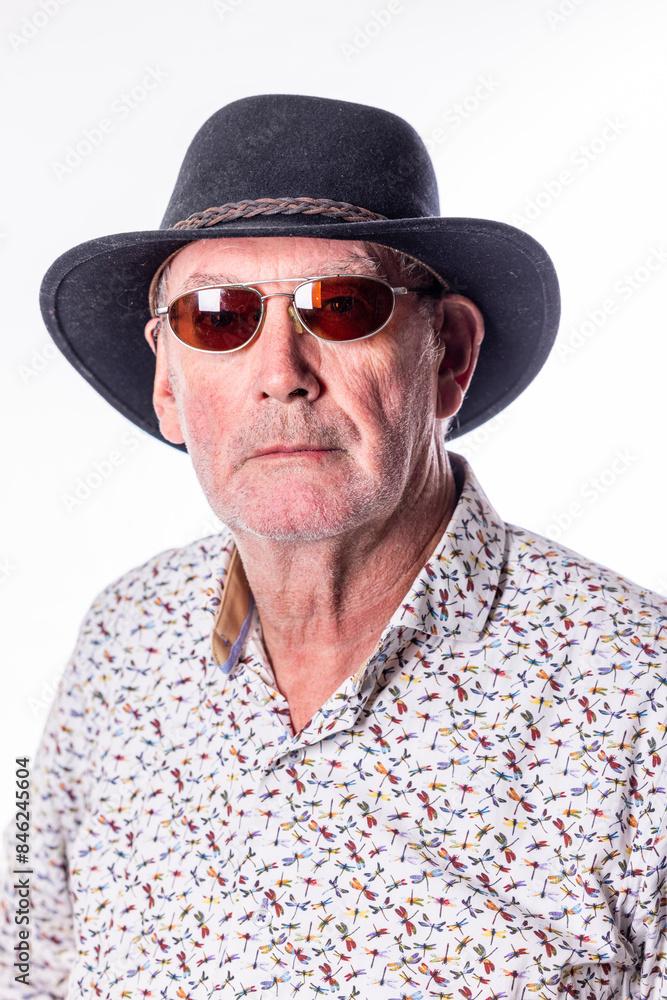 Portrait of an older man wearing a hat and sunglasses, with a patterned shirt, against a white backdrop. Portrait of an older man wearing a hat and sunglasses, with a patterned shirt, against a white backdrop.