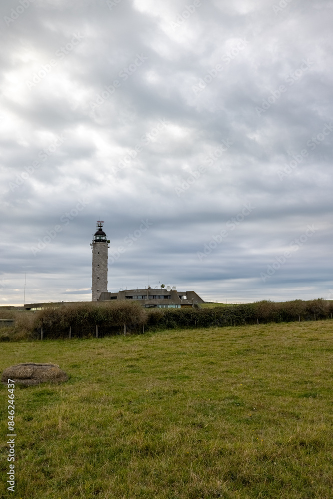 A tall lighthouse stands in a green field under a cloudy sky, surrounded by visible farm structures A tall lighthouse stands in a green field under a cloudy sky, surrounded by visible farm structures