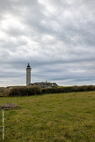 A tall lighthouse stands in a green field under a cloudy sky, surrounded by visible farm structures