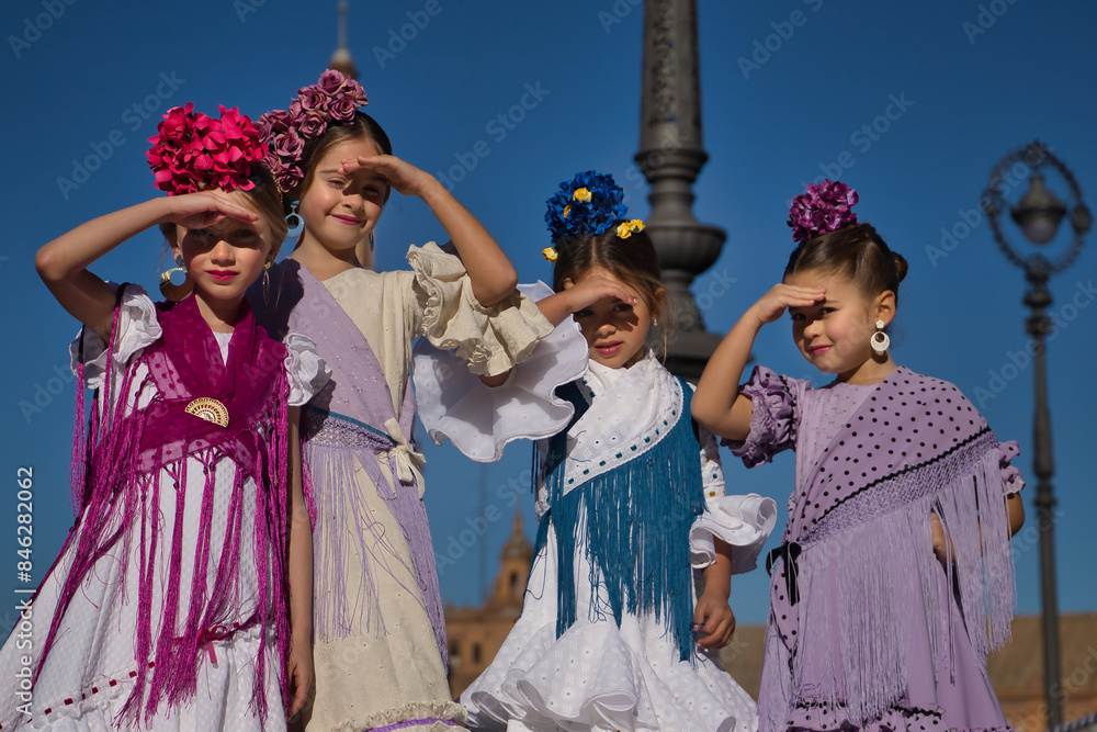 Naklejka premium Four girls dancing flamenco, covering their eyes from the sun with their hands, in typical flamenco costumes on a bridge in a beautiful square in Seville. Dance concept, flamenco, typical Spanish.