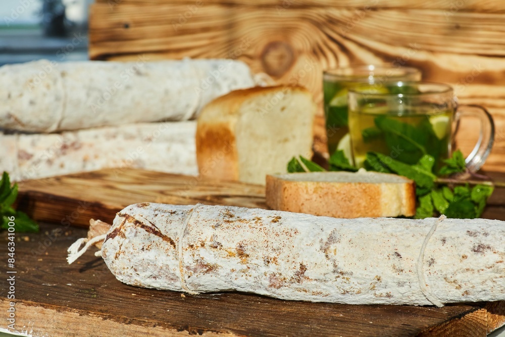 Dried or smoked sausage with white mold on a wooden table. In the background is a loaf of bread and cups of mint-lemon tea. Meat production. Dried or smoked sausage with white mold on a wooden table. In the background is a loaf of bread and cups of mint-lemon tea. Meat production.