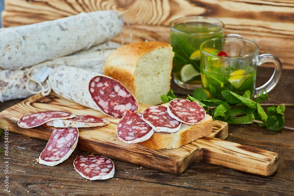 Sliced dried or smoked sausage with white mold on a wooden table. In the background is a loaf of bread and cups of mint-lemon tea. Meat production. Sliced dried or smoked sausage with white mold on a wooden table. In the background is a loaf of bread and cups of mint-lemon tea. Meat production.