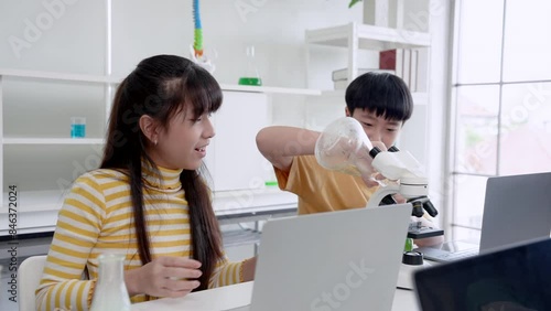 Students' learning in science classroom. Boy in orange shirt concentrates on using microscope, while girl examines flask, in classroom with laptops and scientific glassware on table.