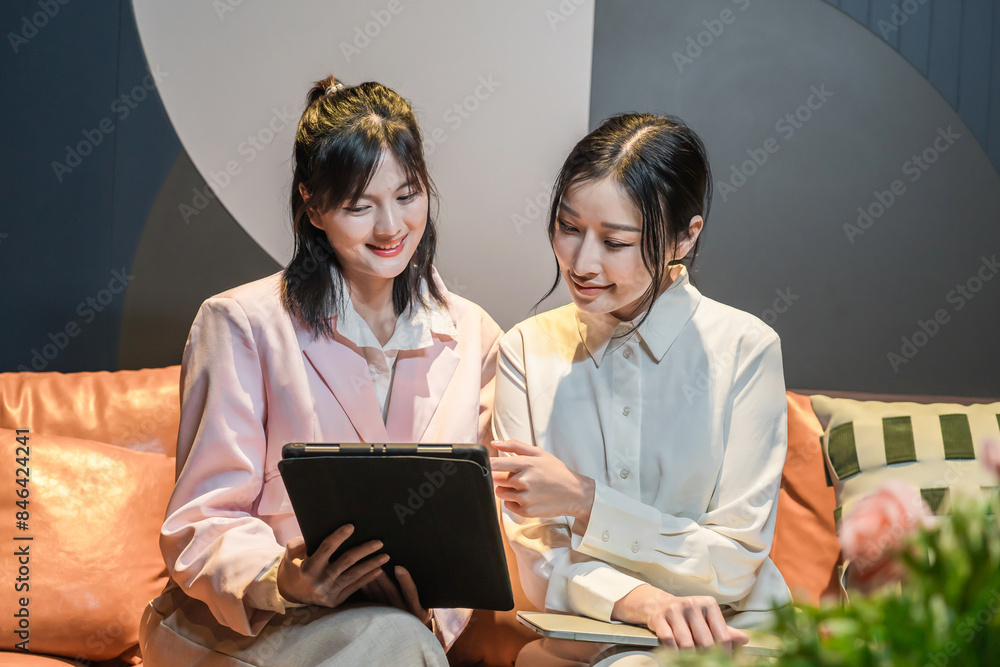 Two happy young asian businesswomen looking at laptops sitting on the sofa in the living room at office in night. and work online with a laptop computer Friends are excited about online shopping. Two happy young asian businesswomen looking at laptops sitting on the sofa in the living room at office in night. and work online with a laptop computer Friends are excited about online shopping.