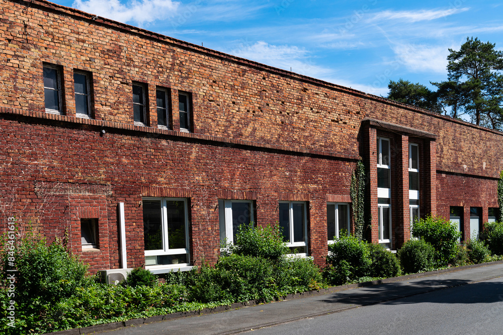 Wall of an old red brick industrial building with overgrown bushes under a blue sky with light white clouds. Wall of an old red brick industrial building with overgrown bushes under a blue sky with light white clouds.