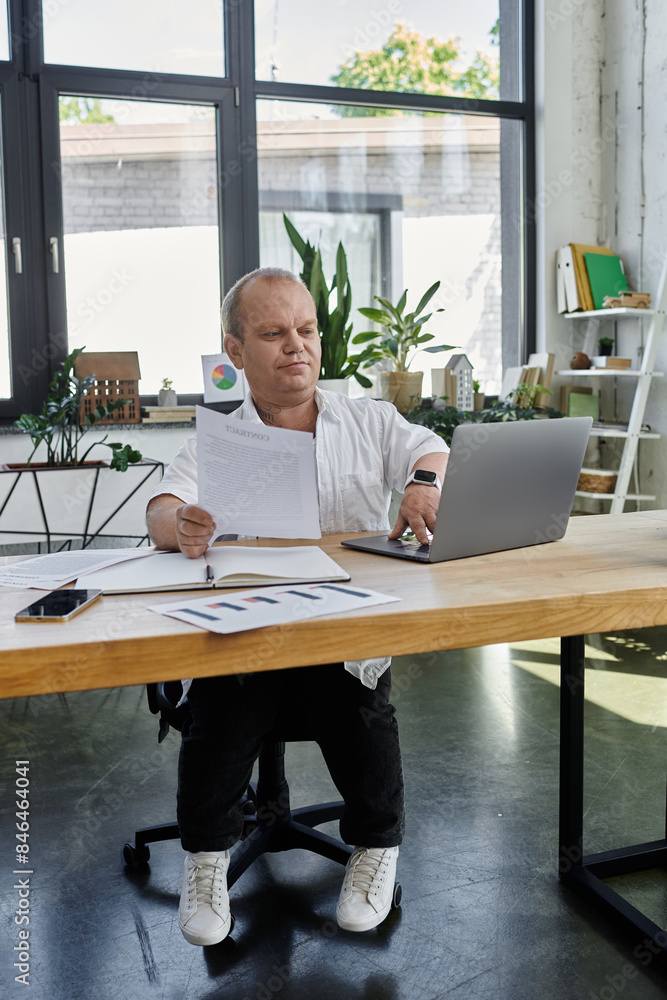A man with inclusivity sits at a desk in a modern office, working on a laptop and reviewing documents. A man with inclusivity sits at a desk in a modern office, working on a laptop and reviewing documents.