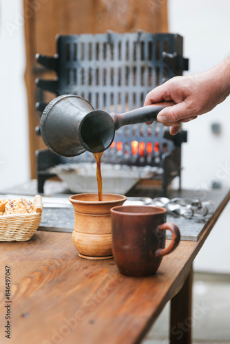A hand pours hot coffee from a jezvah into a clay mug. A jezvah against the background of a brazier, in which fire and smoke are visible.