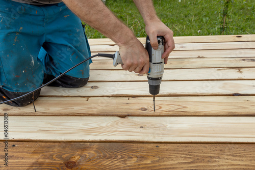 A carpenter kneels on a newly constructed wooden platform, using a cordless drill to screw a fastener into the planks.
