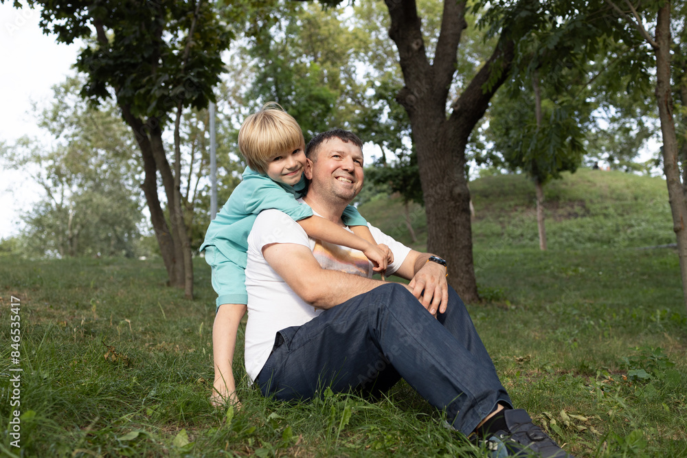 Fototapeta premium Portrait of a happy dad and son. A cute boy 6-7 years old hugs his father sitting on the grass. Walk in the park. Father's Day, Son's Day. Tender relationships in the family