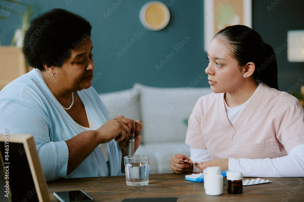 Young nurse holding pills in hands and explaining recommendations while senior African American woman listening carefully Young nurse holding pills in hands and explaining recommendations while senior African American woman listening carefully