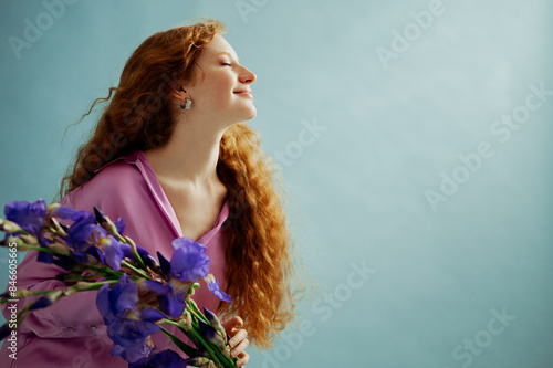 Beautiful happy smiling redhead freckled woman with long curly hair holding bouquet of iris flowers, posing on blue background. Close up studio portrait. Copy, empty, blank space for text