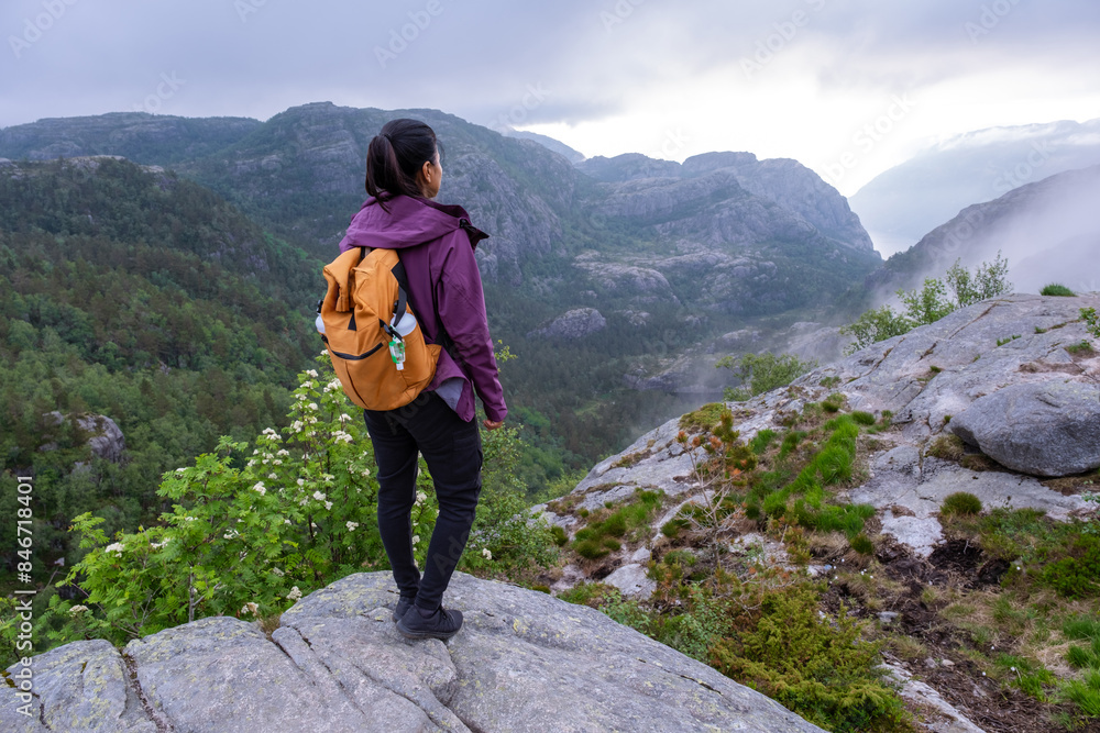 A woman with a backpack stands on a cliffside, gazing at a stunning view of a Norwegian fjord. The mountains are covered in mist, and the landscape is breathtaking. Preikestolen, Norway A woman with a backpack stands on a cliffside, gazing at a stunning view of a Norwegian fjord. The mountains are covered in mist, and the landscape is breathtaking. Preikestolen, Norway