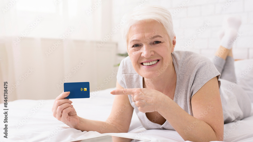 Senior woman is laying on a bed and holding a credit card in her hand. She appears to be focused on the card, possibly making a purchase online or checking her balance Senior woman is laying on a bed and holding a credit card in her hand. She appears to be focused on the card, possibly making a purchase online or checking her balance