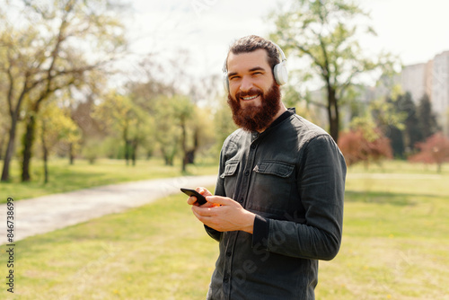 Smiling bearded man using smartphone outdoors in a park on a sunny day, showcasing technology and lifestyle.