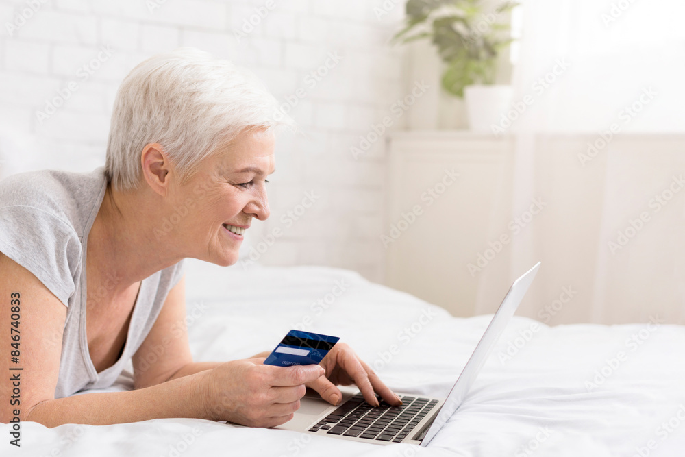 Consumerism and seasonal sales. Senior woman using her credit card and laptop in bed lying in bed, panorama, free space Consumerism and seasonal sales. Senior woman using her credit card and laptop in bed lying in bed, panorama, free space