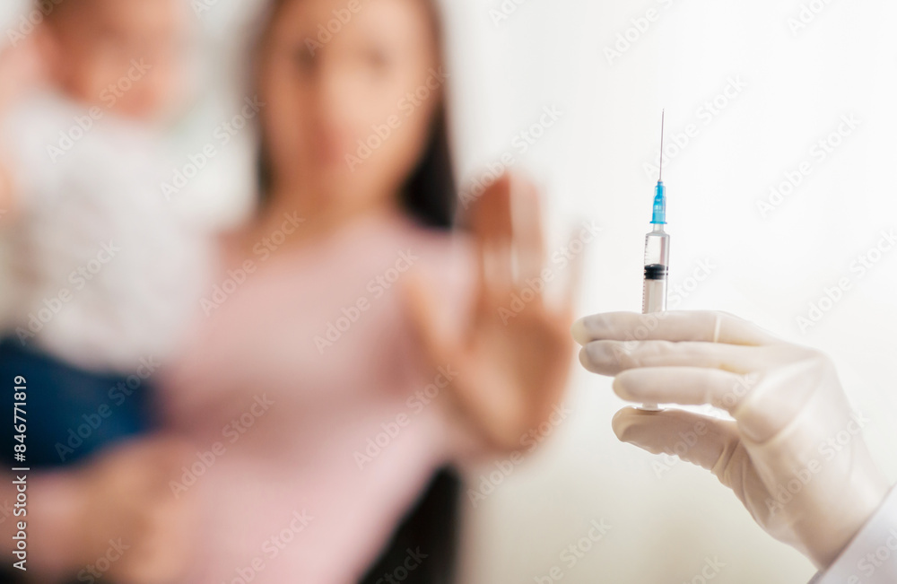 A parent holding a young child raises their hand in refusal as a healthcare worker attempts to administer a vaccination in a clinic setting. A parent holding a young child raises their hand in refusal as a healthcare worker attempts to administer a vaccination in a clinic setting.