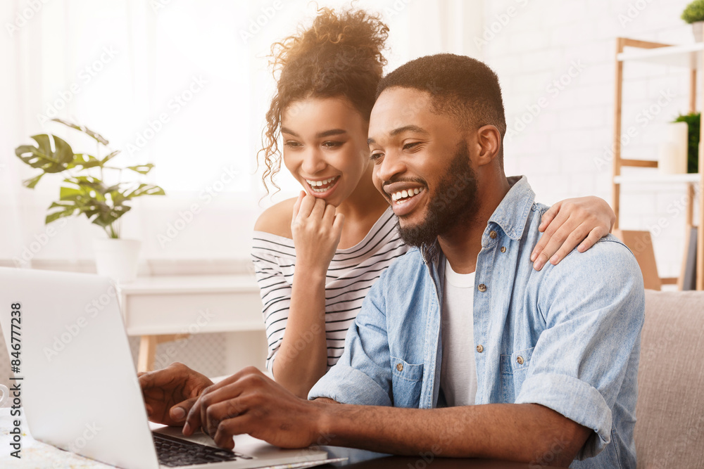 Obraz premium African American man and woman are seated on a couch, both focused on a laptop placed in front of them, engaged in whatever is on the screen, with expressions of concentration and interest.