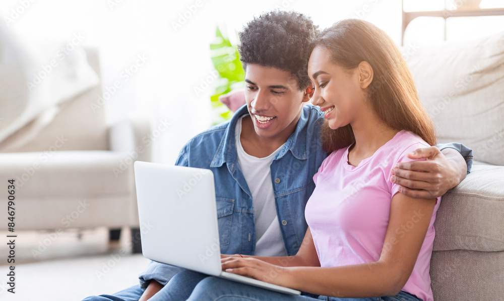 A teen couple sits together on the floor of their living room, using a laptop, both smiling and looking at the screen, watching video content. The guy has his arm around the girls shoulder A teen couple sits together on the floor of their living room, using a laptop, both smiling and looking at the screen, watching video content. The guy has his arm around the girls shoulder