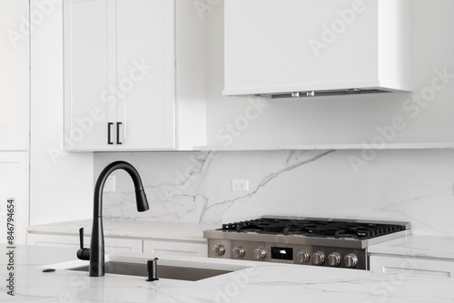 Photos An all white kitchen with a black faucet on a white marble countertop with a stainless steel stove below a white range hood and marble backsplash
