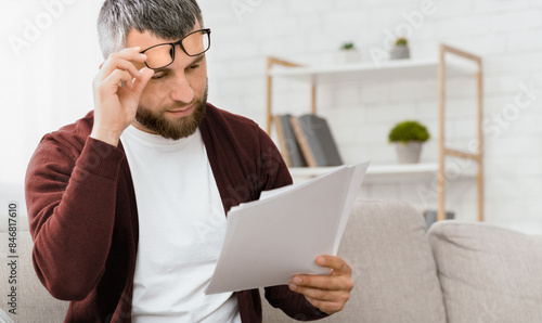 Fotografía A bearded man, wearing glasses and a maroon cardigan, is carefully reading through documents while seated on a light-colored couch in a well-lit, modern living room