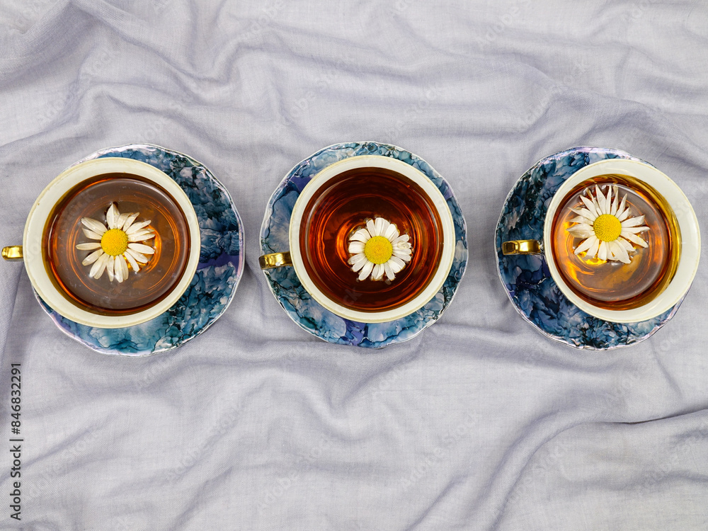Porcelain blue cups with saucers, decorated with gilding, filled with chamomile herbal tea standing in a row on a light blue tablecloth Porcelain blue cups with saucers, decorated with gilding, filled with chamomile herbal tea standing in a row on a light blue tablecloth