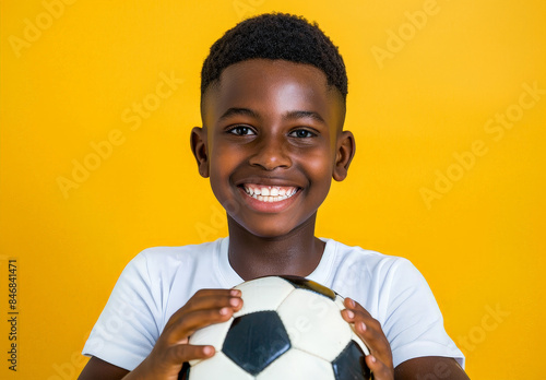 A joyful young black boy stands against a wall, grinning widely as he holds a soccer ball in his hands, dressed in casual clothing for an indoor game