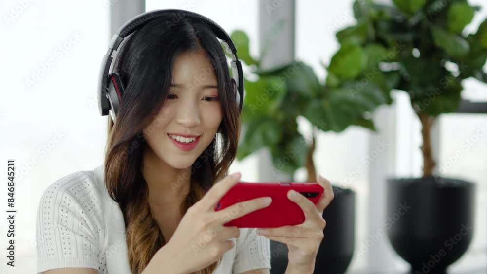Happy young asian woman gamer winner wear headphone competition play video game online with smartphone, excited girl screaming yes winning online game app on cell phone at home Happy young asian woman gamer winner wear headphone competition play video game online with smartphone, excited girl screaming yes winning online game app on cell phone at home