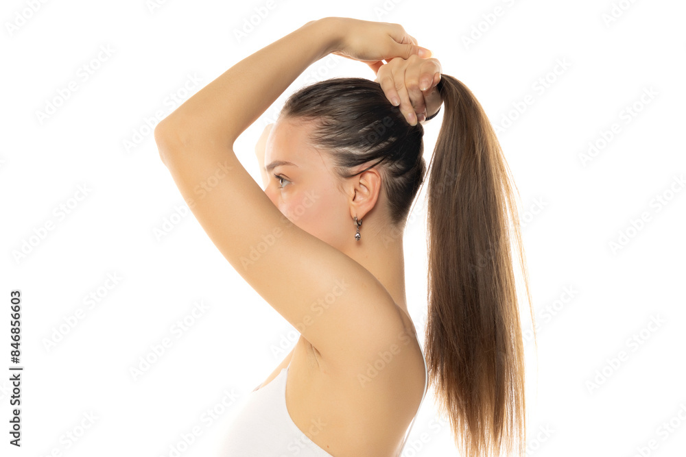 Fototapeta premium Side view of a young modern woman with a partially shaved head tying a ponytail on a white background