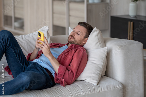 Photography Disinterested man with phone lying on couch surfing online web, procrastination at home