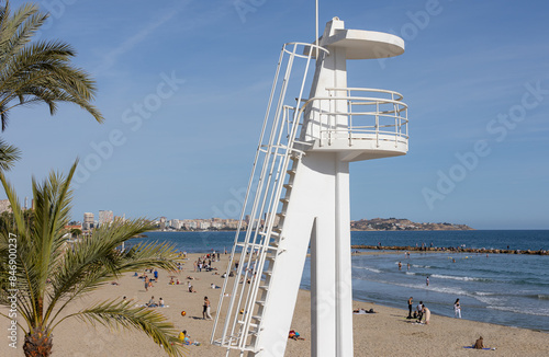 Playa del Postiguet, Alicante, Spain. Landscape of a sandy beach with a lifeguard tower. The Alicante City Beach. Sea beach vacation in low season. Resort spot with people. Swim and sunbath under blue photo