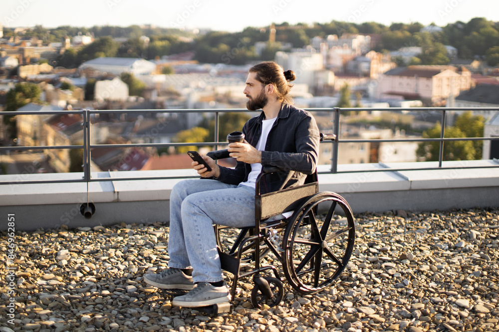 Young man in wheelchair holding smartphone and coffee cup while sitting on rooftop with city view in background. Young man in wheelchair holding smartphone and coffee cup while sitting on rooftop with city view in background.