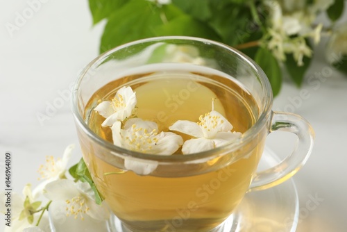 Aromatic jasmine tea in cup, flowers and green leaves on white table, closeup