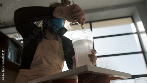 Craftswoman, working in a workshop with toxic materials and wearing a respirator, mixes a viscous, transparent liquid for use in painting. The workshop is flooded with sunlight. Mid shot. Slow motion photo