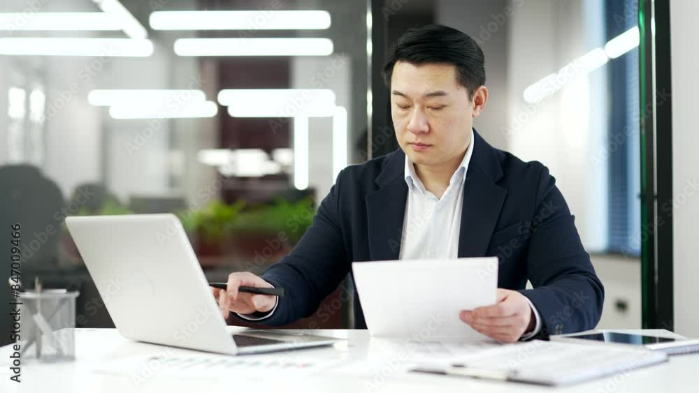 Busy asian businessman is doing paperwork using an application on a laptop sitting at workplace. Confident mature man in a formal suit working fills out documents, writing, banking in business office Busy asian businessman is doing paperwork using an application on a laptop sitting at workplace. Confident mature man in a formal suit working fills out documents, writing, banking in business office